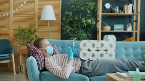 Wanita muda bertopeng wajah menggunakan smartphone di sofa dengan sebungkus tisu toilet di apartemen — Stok Video