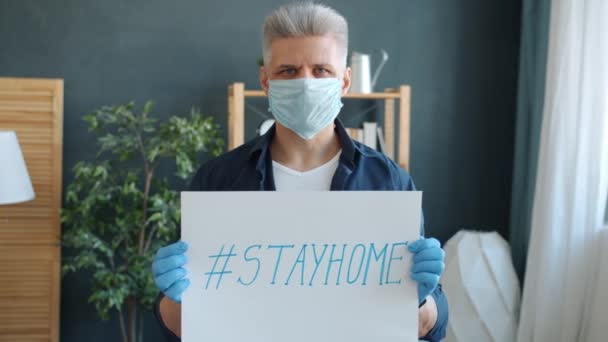 Porträtt av seriös kille i ansiktsmask och handskar som håller stayhome banner i lägenheten — Stockvideo