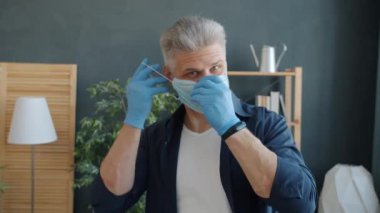 Evde kameraya bakarak tıbbi maske takan yakışıklı bir adam yavaş çekim yapıyor.