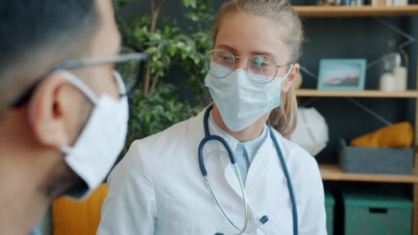 Młoda kobieta lekarz w masce twarzy rozmawia z męskim pacjentem w domu podczas męskiego kanapek — Wideo stockowe