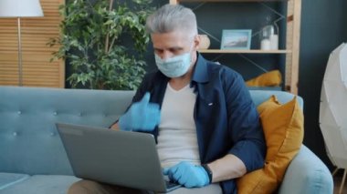 Tıbbi maskeli ve eldivenli bir adam laptopta öksürüyor ve konuşuyor. Evden video görüşmesi yapıyor.