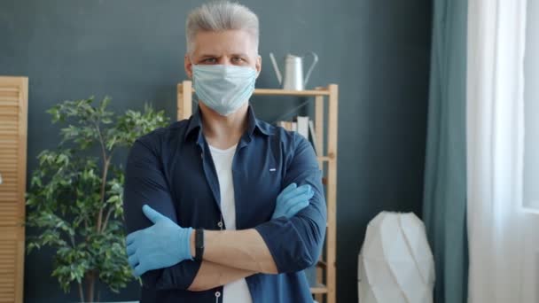 Portret dorosłego mężczyzny w zapobiegawczej masce medycznej i rękawiczkach stojących w mieszkaniu z skrzyżowanymi ramionami — Wideo stockowe