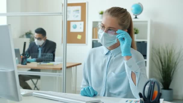 Ragazza bionda in maschera e guanti fare videochiamata online con il computer al lavoro parlando gesticolando — Video Stock