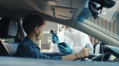 Quarantive Kostüm giyen adam dışarıda ateşini ölçen kadın sürücüyle konuşuyor.
