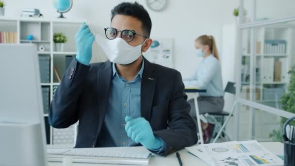 Portret van een Arabier die een medisch masker afdoet zittend aan een bureau terwijl hij naar de camera kijkt — Stockvideo