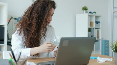 Genç bayan ofis çalışanı dezenfektan kullanıyor, ellerini siliyor sonra da iş yerinde dizüstü bilgisayarla çalışıyor.