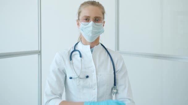 Movimento lento del giovane medico in abito bianco, maschera facciale e occhiali incrociando le braccia in clinica — Video Stock