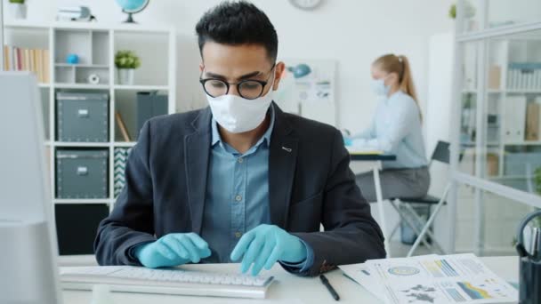 Hombre de Oriente Medio con máscara y guantes trabajando con computadora en la oficina durante el encierro — Vídeos de Stock