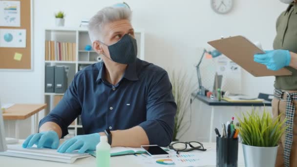 Человек в маске занят компьютерной работой, пока сотрудник измеряет температуру инфракрасным термометром — стоковое видео