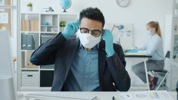 Retrato del hombre árabe poniéndose máscara médica facial en la oficina durante la epidemia — Vídeos de Stock