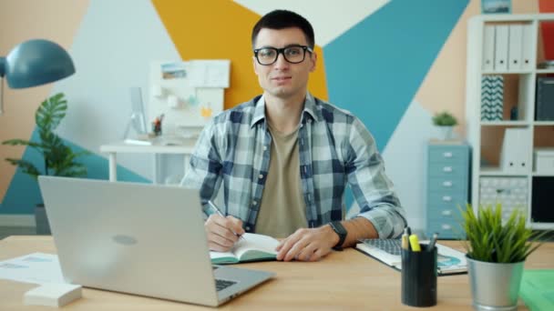 Портрет молодого человека в очках, смотрящего в камеру, сидящего за столом с ручкой и блокнотом — стоковое видео
