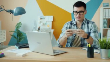 Modern ofiste gözlüklü mutlu adam akıllı telefonuyla video oyunu oynuyor.