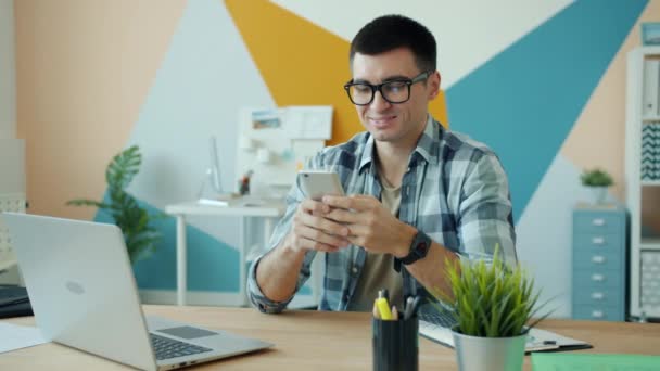 Knappe jongeman met smartphone op kantoor aan de balie genieten van online communicatie — Stockvideo
