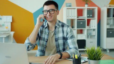 Gülümseyen adam ofis çalışanı cep telefonuyla konuşuyor modern işyerinde masa başında çalışıyor.