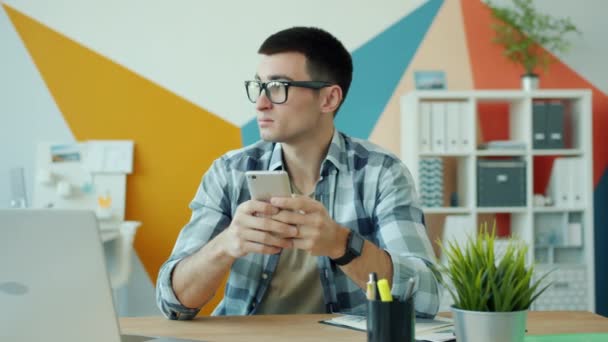 Vrolijke man met behulp van smartphone sms 'en en glimlachen aan het bureau op het werk — Stockvideo