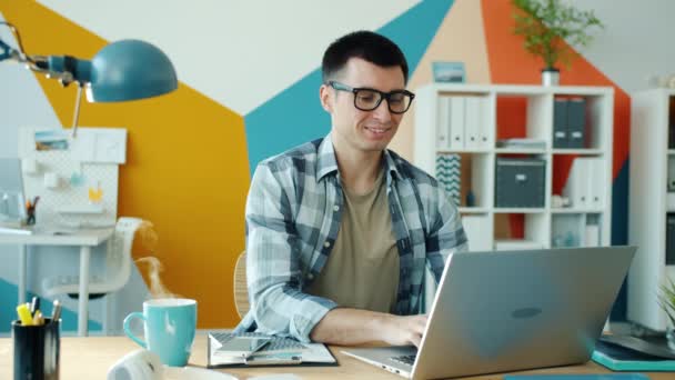 Нескінченна петля людини, що працює з ноутбуком і посміхається, коли пара піднімається з чашки кави в офісі — стокове відео