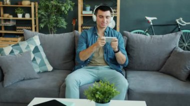 Kulaklıklı neşeli adam evde akıllı telefon kullanarak video oyunu oynuyor.