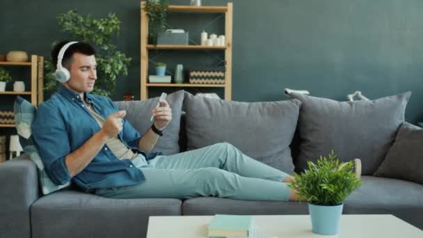 Langzame beweging van vrolijke man in hoofdtelefoons luisteren naar muziek kijken smartphone scherm — Stockvideo