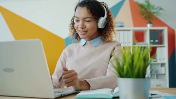 Chica alegre trabajador de oficina que trabaja con el ordenador portátil y escuchar música a través de auriculares en el trabajo — Vídeos de Stock