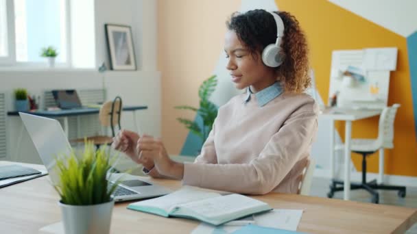 Verspielte Afroamerikanerin, die mit Laptop im Büro arbeitet und Kopfhörer trägt und Spaß hat — Stockvideo