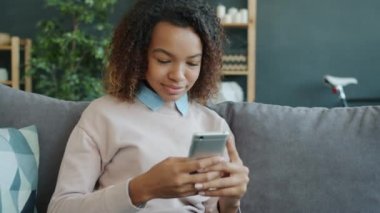 Çekici Afro-Amerikalı kız elinde akıllı telefonla ekranlara dokunuyor evde dinleniyor