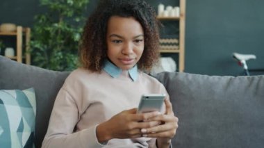 Genç Afro-Amerikalı kadın modern apartmandaki akıllı telefondan sosyal medyadan keyif alıyor.