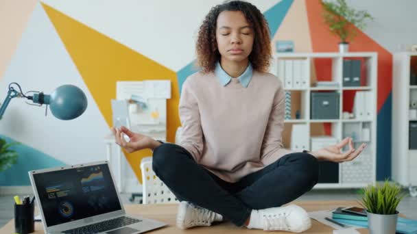 Молодая афроамериканская девушка сидит в позе лотоса на офисном столе, наслаждаясь отдыхом и медитацией — стоковое видео