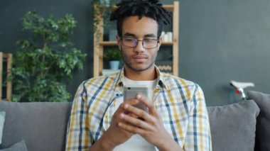 Mutlu Afrikalı Amerikalı öğrenci evde akıllı telefon kullanıyor. Kanepede yalnız başına dinleniyor.