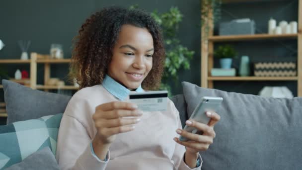 Movimento lento de mulher de raça mista feliz pagando com cartão de crédito e smartphone em casa — Vídeo de Stock