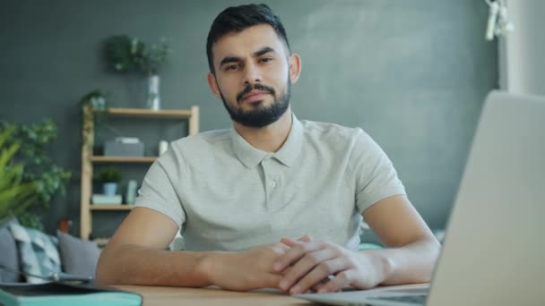 Portret van een aantrekkelijke jonge Arabische man kijkend naar camera zittend aan tafel met laptop thuis — Stockvideo
