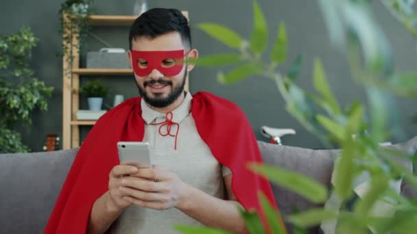 Succesvolle man in super held kostuum met behulp van smartphone glimlachen op de bank in appartement — Stockvideo