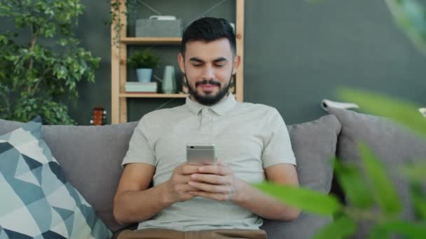 Hombre árabe despreocupado utilizando mensajes de texto de teléfonos inteligentes de navegación en el interior en el sofá en casa — Vídeo de stock