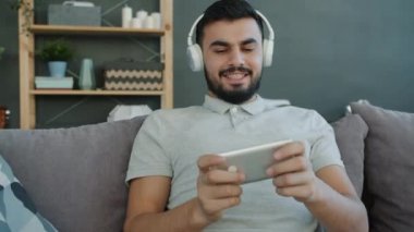 Neşeli Arap adam kulaklıkla müzik dinlemekten ve akıllı telefondan video oyunu oynamaktan hoşlanıyor.
