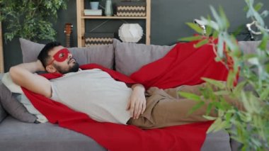 Süper kahraman kostümlü, kırmızı pelerinli ve maskeli bir adam evdeki kanepede uyuyor.