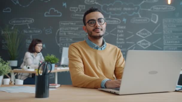 Portrét seriózního mladého Araba s laptopem sedícím na pracovišti v otevřené kanceláři — Stock video