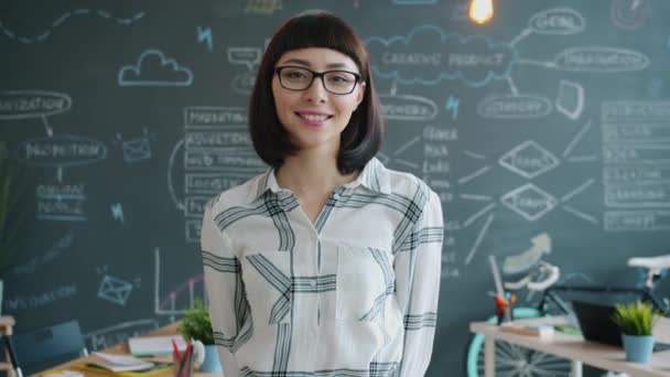 Retrato de linda chica de pie en la oficina creativa y sonriendo mirando a la cámara — Vídeos de Stock