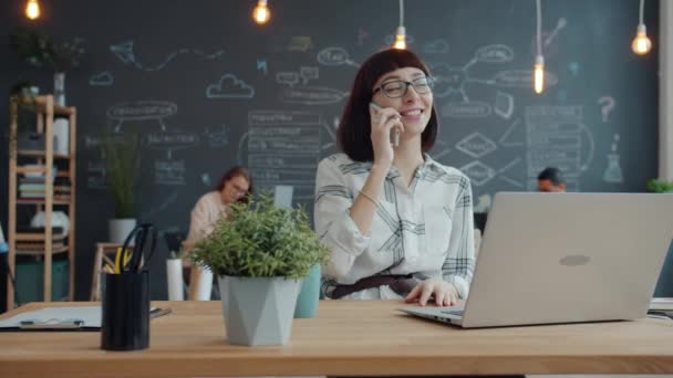 Iloinen tyttö puhuu matkapuhelimeen ja nauraa työpöydälle avoimessa tilassa toimistossa — kuvapankkivideo