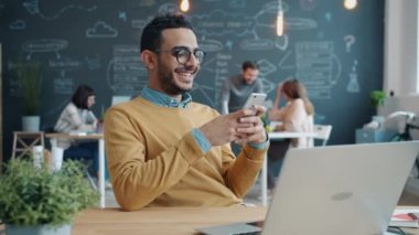 Neşeli bir Arap akıllı telefon kullanıyor. İnternetten mesajlaşıyor ve ofiste sırıtıyor.