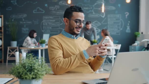 Glücklicher Araber mit Smartphone und Lächeln am Schreibtisch in gemeinsamem Arbeitsplatz — Stockvideo