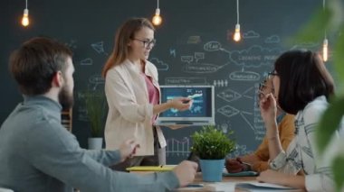 Kız ofis çalışanı sunum yapıyor. Dizüstü bilgisayara işaret ederek iş arkadaşlarıyla konuşuyor.