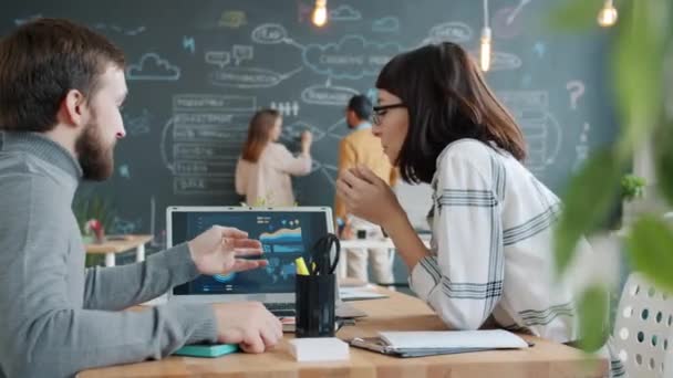 Chica y chico hablando mirando la pantalla del ordenador portátil mientras sus colegas escriben en pizarra en la oficina — Vídeos de Stock