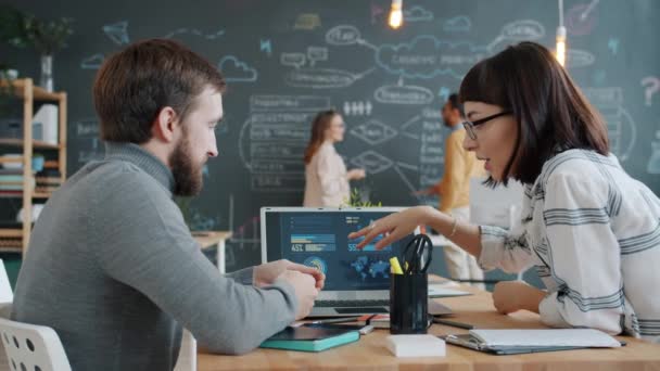 Colegas falando sobre negócios usando laptop enquanto homem e mulher escrevendo no quadro no fundo — Vídeo de Stock