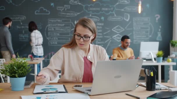 Junge Frau arbeitet mit Laptop im Büro, während Kollegen im Hintergrund auf Kreide schreiben — Stockvideo