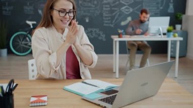 Heyecanlı ofis çalışanı el çırpıyor ve gülümsüyor paylaşılan ofiste dizüstü bilgisayara bakıyor.