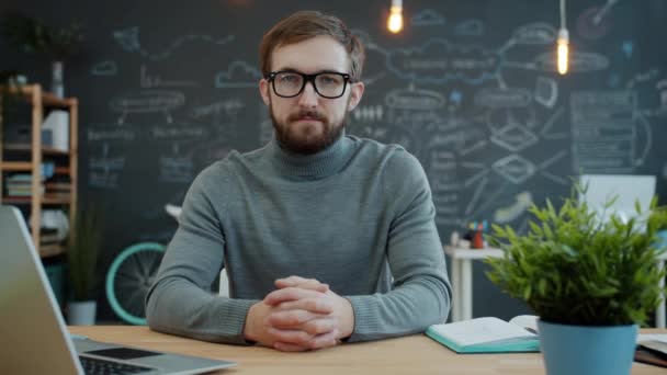 Portré fiatal férfi szemüvegben nézi kamera ül az asztalnál, laptop számítógép látható — Stock videók