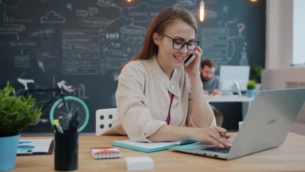 Blond flicka som arbetar med bärbar dator i kontor skriva och tala på mobiltelefon — Stockvideo