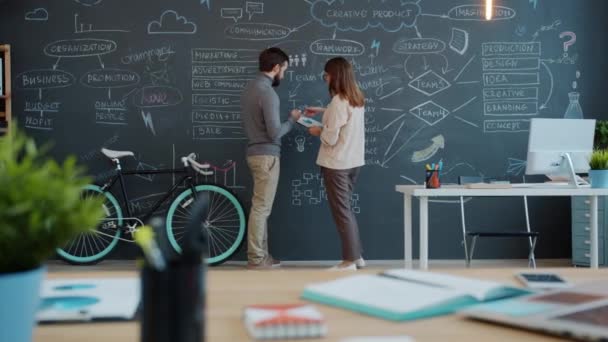 Girl and guy talking and writing on blackboard wall in shared office planning business strategy — Stock video