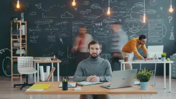 Zoom-in time lapse porträtt av attraktiv ung man vid skrivbordet i delat kontor full av människor — Stockvideo