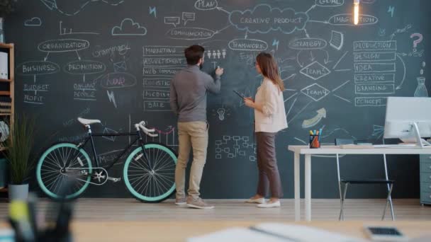 Mann und Frau schreiben auf Kreidetafel und diskutieren Geschäftsstrategie im Großraumbüro — Stockvideo
