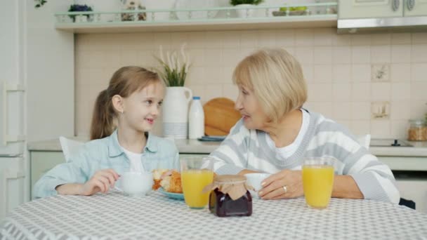 Bucuroasă nepoată de familie și bunică luând micul dejun acasă vorbind râzând — Videoclip de stoc